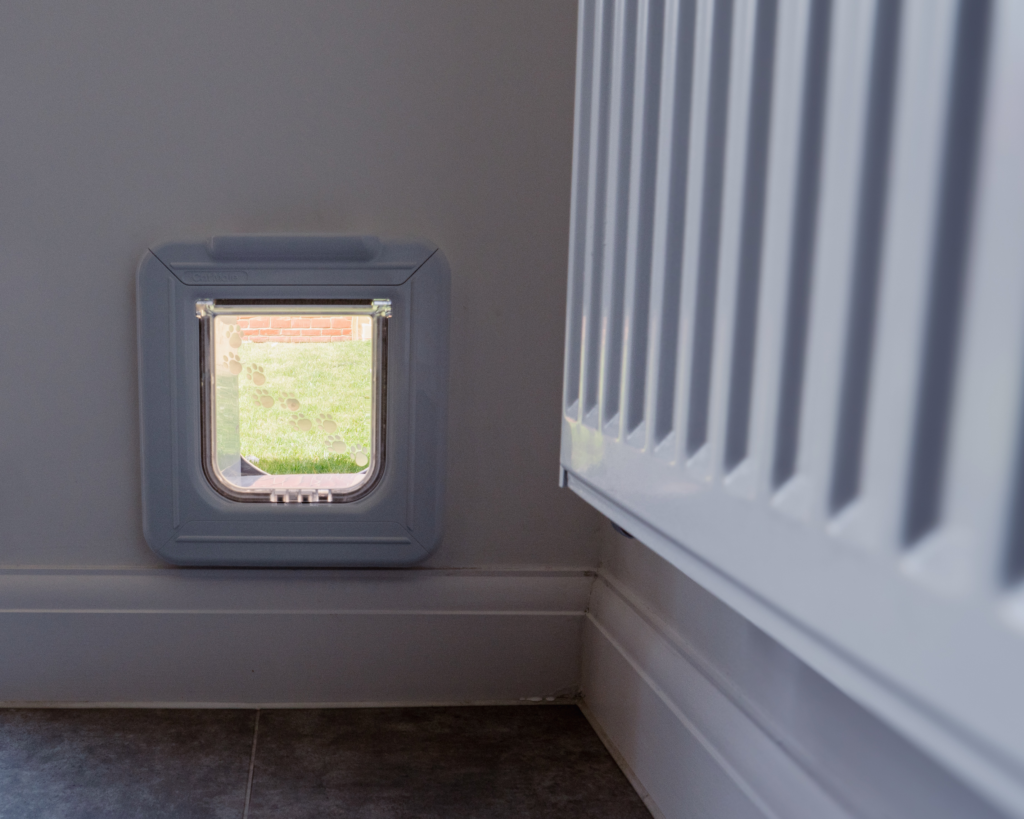 "White modern radiator leading your eye to a White Pet Safe Superdeluxe microchip-activated cat flap, fitted into a grey painted internal wall, leading into the garden. As seen through the clear flap, the interior is bright and inviting, creating a serene atmosphere for pets and their owners". This design choice enhances the overall aesthetic of the space, making it both functional and visually appealing.