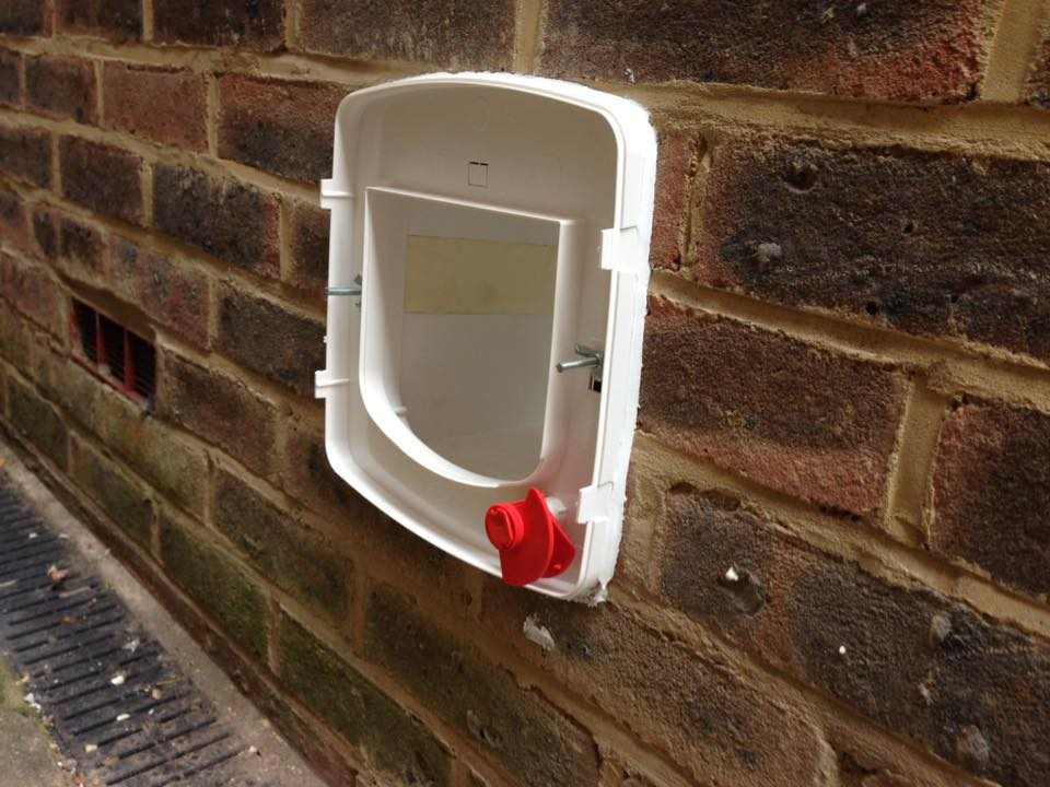 white cat flap with red knob fitted to a weathered, brick wall